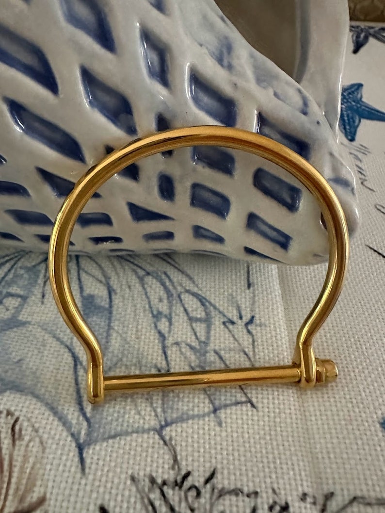 May include: A gold-colored, hinged bracelet with a distinctive U-shaped design. The bracelet features a straight bar that connects to the curved top, creating a sleek, modern aesthetic. The bracelet is displayed on a patterned surface.