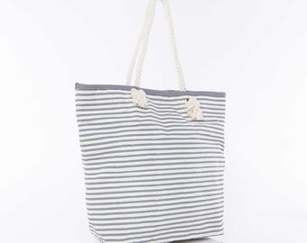 May include: A gray and white striped tote bag with rope handles and white tassels. The bag is made of canvas and has a relaxed, casual style.