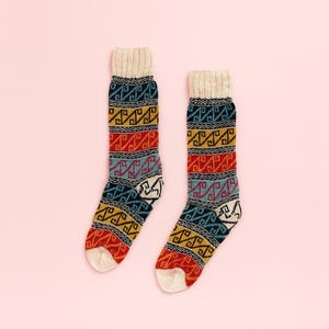 Può includere: Un paio di calzini fantasia con polsino e punta color crema. I calzini presentano un design geometrico in rosso, giallo, blu e bianco. I calzini sono presentati su uno sfondo rosa chiaro.