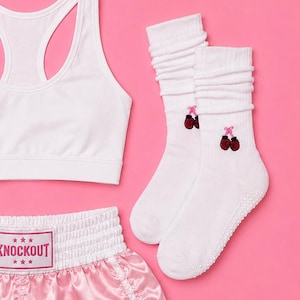 May include: A set of athletic gear including a pair of light pink boxing gloves, a white racerback sports bra, light pink satin boxing shorts with a white waistband featuring the text KNOCKOUT, and white crew socks with small boxing glove icons.