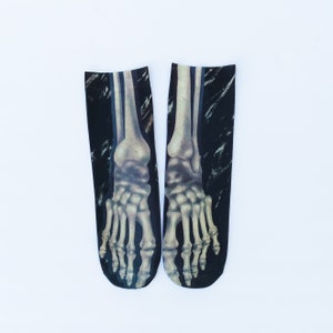 May include: A pair of black crew socks featuring a realistic, detailed print of human foot and ankle bones in shades of off-white and grey. The skeletal design extends from the toe area up to the ankle, set against a dark, textured background.