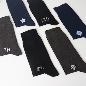 May include: Assortment of personalized socks in various colors, including navy, black, and gray. Each sock features embroidered initials in white thread. Some socks have initials within a star or diamond shape.