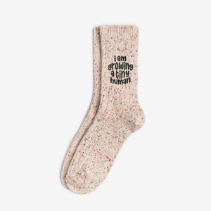Comfortable socks featuring a witty phrase about pregnancy, capturing the transformative experience of growing a tiny human. Cream speckled ribbed socks with colorful flecks and black embroidery reading “I am growing a tiny human.” Neutral maternity socks by Sockmate for pregnancy comfort.