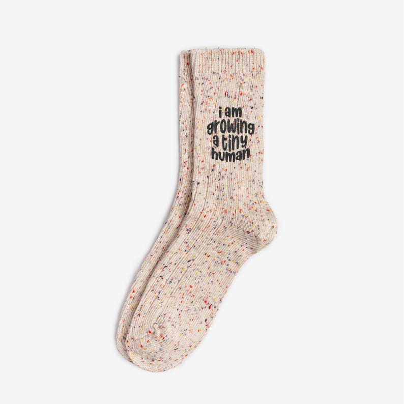 Comfortable socks featuring a witty phrase about pregnancy, capturing the transformative experience of growing a tiny human. Cream speckled ribbed socks with colorful flecks and black embroidery reading “I am growing a tiny human.” Neutral maternity socks by Sockmate for pregnancy comfort.
