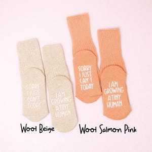 Pregnancy announcement socks in beige and peach with white lettering, photographed on a soft pink background.