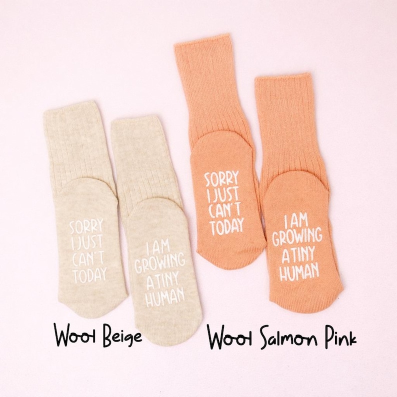 Pregnancy announcement socks in beige and peach with white lettering, photographed on a soft pink background.