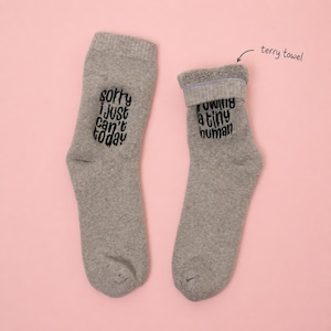 Model wearing gray terry towel socks with white embroidery that reads “Sorry I just can’t today” and “I am growing a tiny human.” Cozy maternity socks set by Sockmate on pink background.