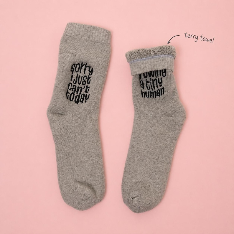 Model wearing gray terry towel socks with white embroidery that reads “Sorry I just can’t today” and “I am growing a tiny human.” Cozy maternity socks set by Sockmate on pink background.