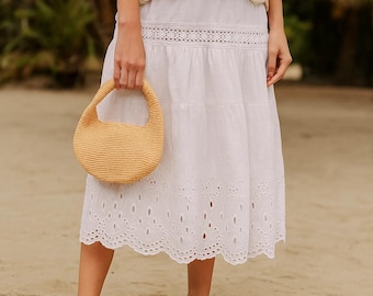 May include: A light yellow woven handbag with a rounded shape and a short handle. The bag is held in front of a white eyelet skirt. The skirt has a scalloped hemline and a decorative band at the waist. The background is a sandy beach.