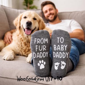 K&ouml;nnte beinhalten: Graue Socken mit dem wei&szlig;en Text "FROM FUR DADDY" und "TO BABY DADDY". Die Socken zeigen einen Pfotenabdruck und Babyf&uuml;&szlig;e. Ein Golden Retriever und eine Person sind im Hintergrund.