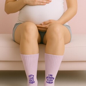 Pregnant woman sitting on pink couch wearing matching pink socks embroidered with “Sorry I just can’t today” and “I am growing a tiny human.” Maternity socks set by Sockmate.
