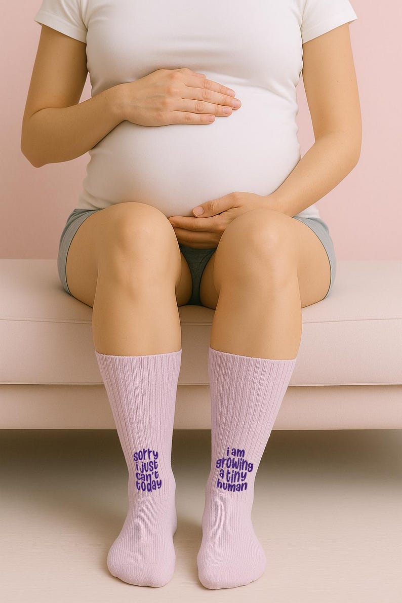 Pregnant woman sitting on pink couch wearing matching pink socks embroidered with “Sorry I just can’t today” and “I am growing a tiny human.” Maternity socks set by Sockmate.