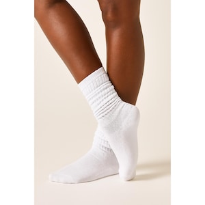 May include: A pair of white, slouch socks. The socks are made of a soft material and have a ribbed texture on the upper part. The socks are worn on a person's feet, with one foot slightly in front of the other.