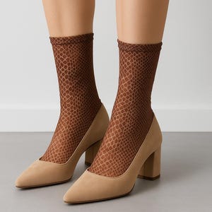 May include: A pair of tan suede pumps with a pointed toe and block heel. The shoes are paired with brown patterned socks. The socks have a reptile-like design. The background is a light gray.
