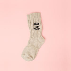 May include: A pair of beige ribbed knit socks with the text "SHHHH, I am reading" in black. The socks are on a light pink surface. These socks are a comfortable accessory for reading.