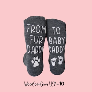 K&ouml;nnte beinhalten: Ein Paar dunkelgraue Socken mit wei&szlig;em Text und Grafiken. Die linke Socke tr&auml;gt die Aufschrift "FROM FUR DADDY" mit einem Pfotenabdruck, und die rechte Socke tr&auml;gt die Aufschrift "TO BABY DADDY" mit Babyf&uuml;&szlig;en.