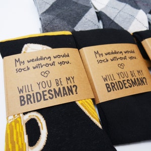 Könnte beinhalten: Zwei Paar Socken mit einer braunen Karte, die an jedem befestigt ist. Die Karte trägt den Text "My wedding would sock without you." und "Will you be my Bridesman?" darauf. Ein Paar Socken hat ein gelbes und schwarzes Design mit einem Bierkrug darauf. Das andere Paar Socken hat ein schwarzes und graues Argyle-Muster.