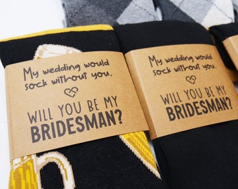 May include: Two pairs of socks with a brown card attached to each. The card has the text "My wedding would sock without you." and "Will you be my Bridesman?" on it. One pair of socks has a yellow and black design with a beer mug on it. The other pair of socks has a black and grey argyle pattern.