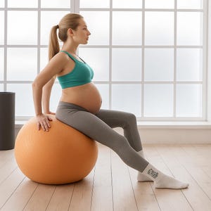 Pregnant woman sitting on orange yoga ball in a bright studio wearing white pregnancy socks with motivational text. Comfortable prenatal socks ideal for pilates or yoga.