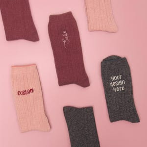 Könnte beinhalten: Auswahl an gerippten Socken in verschiedenen Farben, darunter Burgunderrot, Grau und Rosa. Einige Socken haben gestickten Text: "Custom", "Selin" und "your design here". Die Socken sind auf einem rosa Hintergrund angeordnet.