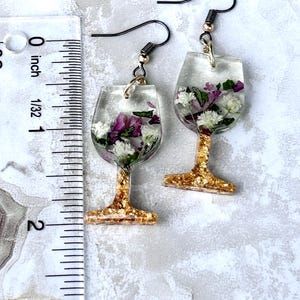 May include: A pair of gold-rimmed glass wine glass earrings with dried flowers encased in clear resin. The earrings are approximately 1 inch long and have black metal hooks.