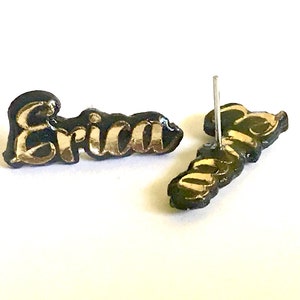 May include: A pair of stud earrings spelling the name "Erica" in gold lettering on a black background. The earrings are in a cursive font and have a post backing.