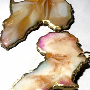 Op de afbeelding: Een paar gouden oorringen in de vorm van het Afrikaanse continent. De oorringen zijn gemaakt van hars en hebben een wit en gouden marmerpatroon. Eén oorring heeft een roze rand, terwijl de andere een gouden rand heeft.