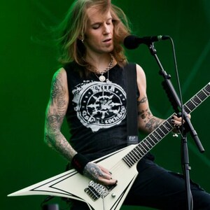 Puede incluir: Un m&uacute;sico tocando una guitarra el&eacute;ctrica blanca con detalles negros en un escenario. El m&uacute;sico lleva una camiseta sin mangas negra con un dise&ntilde;o gr&aacute;fico, pantalones negros y tiene tatuajes. Un micr&oacute;fono est&aacute; posicionado frente a &eacute;l.