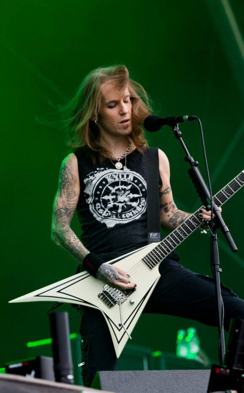 Puede incluir: Un m&uacute;sico tocando una guitarra el&eacute;ctrica blanca con detalles negros en un escenario. El m&uacute;sico lleva una camiseta sin mangas negra con un dise&ntilde;o gr&aacute;fico, pantalones negros y tiene tatuajes. Un micr&oacute;fono est&aacute; posicionado frente a &eacute;l.