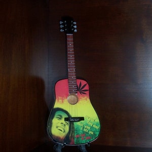 Puede incluir: Una peque&ntilde;a guitarra ac&uacute;stica colorida con un dise&ntilde;o verde, amarillo y rojo. La guitarra tiene una imagen de Bob Marley en la parte delantera con la cita "Lo &uacute;nico de la m&uacute;sica, cuando te golpea, no sientes dolor".