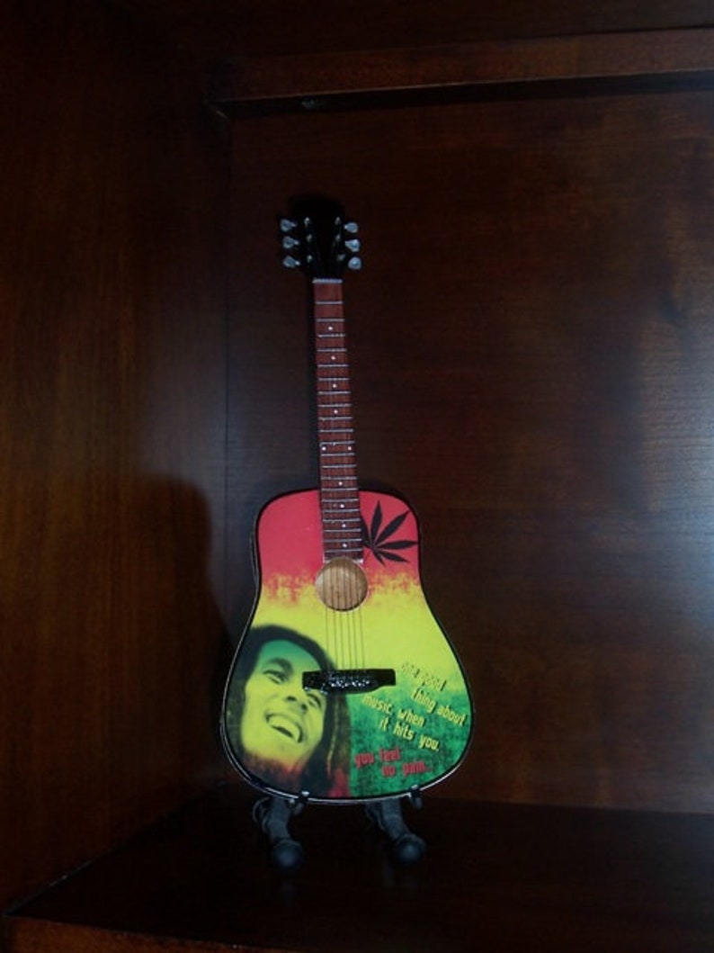 Puede incluir: Una peque&ntilde;a guitarra ac&uacute;stica colorida con un dise&ntilde;o verde, amarillo y rojo. La guitarra tiene una imagen de Bob Marley en la parte delantera con la cita "Lo &uacute;nico de la m&uacute;sica, cuando te golpea, no sientes dolor".