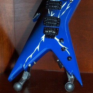 Puede incluir: Una guitarra el&eacute;ctrica azul con dise&ntilde;os de rayos blancos. La guitarra est&aacute; en un soporte y tiene una placa de protecci&oacute;n negra y botones negros.