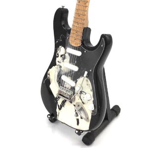 Puede incluir: Una guitarra el&eacute;ctrica negra con m&aacute;stil y diapas&oacute;n de color claro. La guitarra presenta una imagen en blanco y negro en el cuerpo. Se exhibe sobre un soporte negro. La guitarra tiene perillas blancas y herrajes plateados.