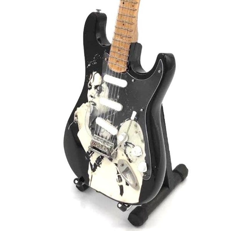 Puede incluir: Una guitarra el&eacute;ctrica negra con m&aacute;stil y diapas&oacute;n de color claro. La guitarra presenta una imagen en blanco y negro en el cuerpo. Se exhibe sobre un soporte negro. La guitarra tiene perillas blancas y herrajes plateados.