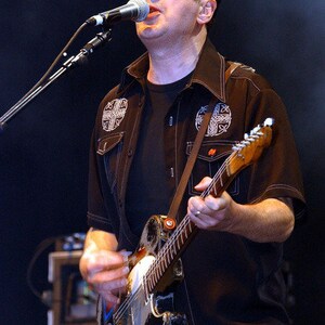 May include: A man in a black shirt with embroidered designs plays a black electric guitar with a white pickguard. He is singing into a microphone.