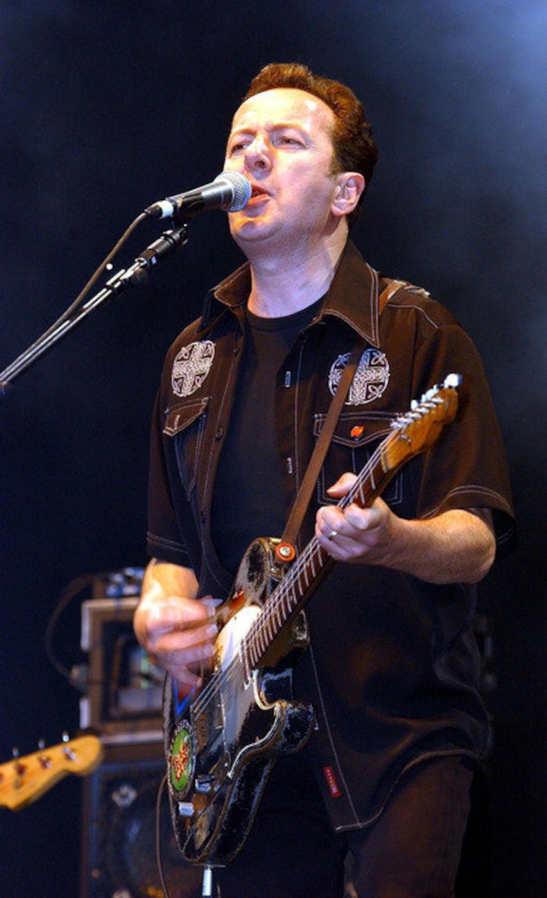 May include: A man in a black shirt with embroidered designs plays a black electric guitar with a white pickguard. He is singing into a microphone.