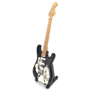 Puede incluir: Guitarra el&eacute;ctrica negra con m&aacute;stil y clavijero de madera clara. El cuerpo de la guitarra presenta un dise&ntilde;o gr&aacute;fico en blanco y negro. La guitarra se exhibe sobre un soporte negro sobre fondo blanco.
