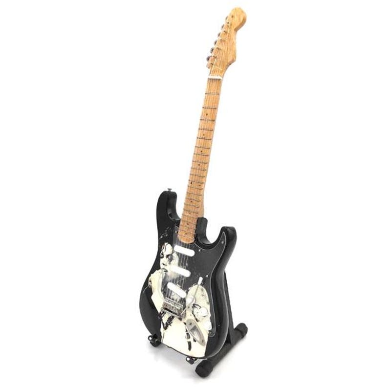 Puede incluir: Guitarra el&eacute;ctrica negra con m&aacute;stil y clavijero de madera clara. El cuerpo de la guitarra presenta un dise&ntilde;o gr&aacute;fico en blanco y negro. La guitarra se exhibe sobre un soporte negro sobre fondo blanco.