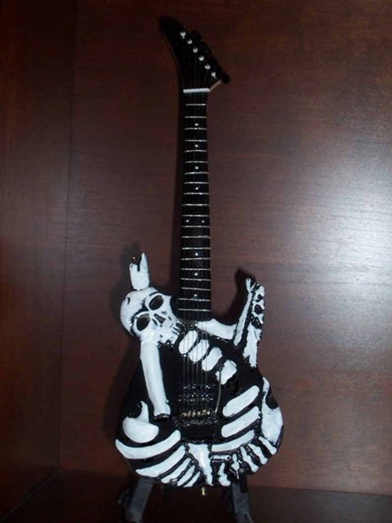 May include: A black electric guitar with a white skeleton design. The guitar has a black body and a black neck with white fret markers. The skeleton design is painted on the body of the guitar and includes a skull and crossbones.