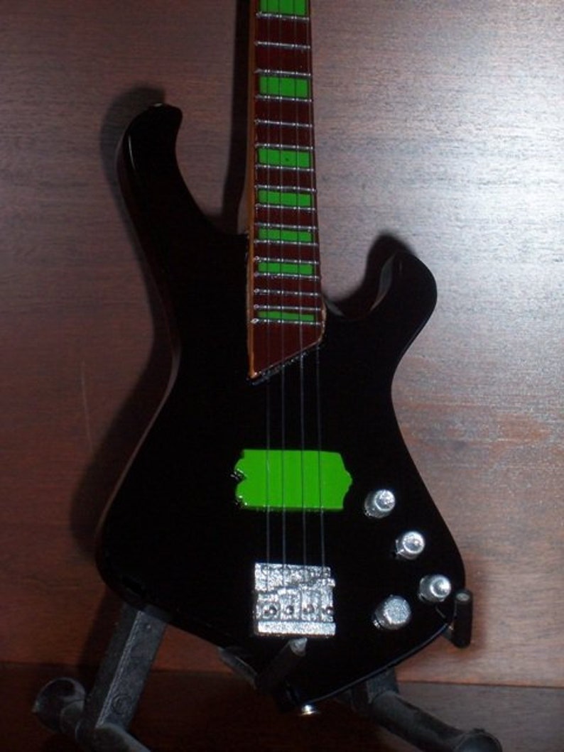 Puede incluir: Un bajo el&eacute;ctrico negro con un diapas&oacute;n rayado verde y marr&oacute;n. La guitarra tiene una forma de cuerpo &uacute;nica y est&aacute; en un soporte.