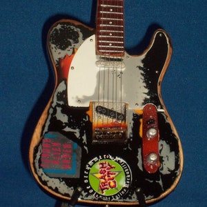 May include: A black and white electric guitar with a white pickguard and a green, yellow, and red sticker that says "Trash City" on it. The guitar has a wooden body and a silver metal bridge.