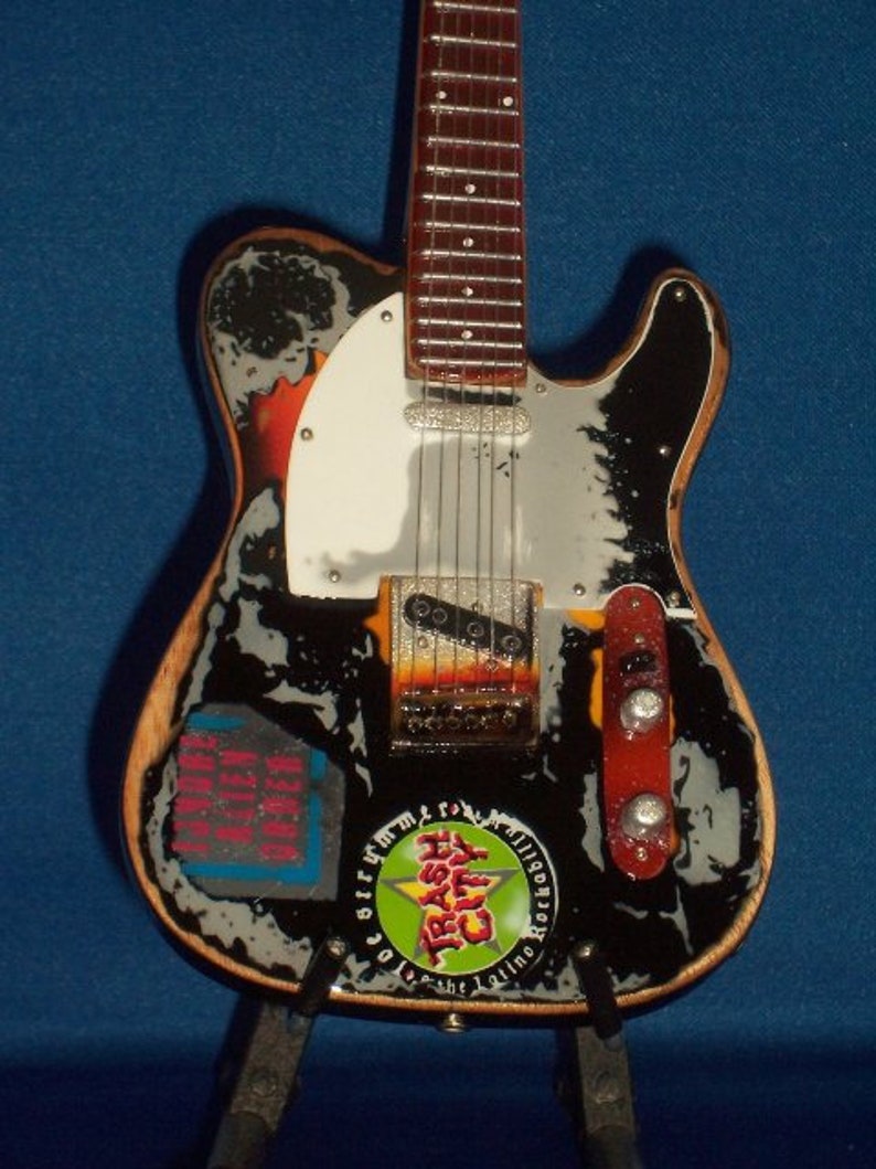 May include: A black and white electric guitar with a white pickguard and a green, yellow, and red sticker that says "Trash City" on it. The guitar has a wooden body and a silver metal bridge.