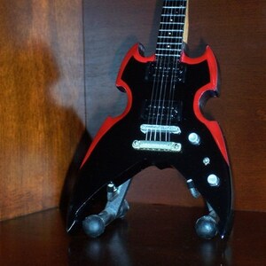 Puede incluir: Una guitarra el&eacute;ctrica negra con detalles rojos. La guitarra tiene una forma &uacute;nica con una pala puntiaguda y un cuerpo que se asemeja a las alas de un murci&eacute;lago. La guitarra tiene una sola pastilla y un puente con una barra de tr&eacute;molo.