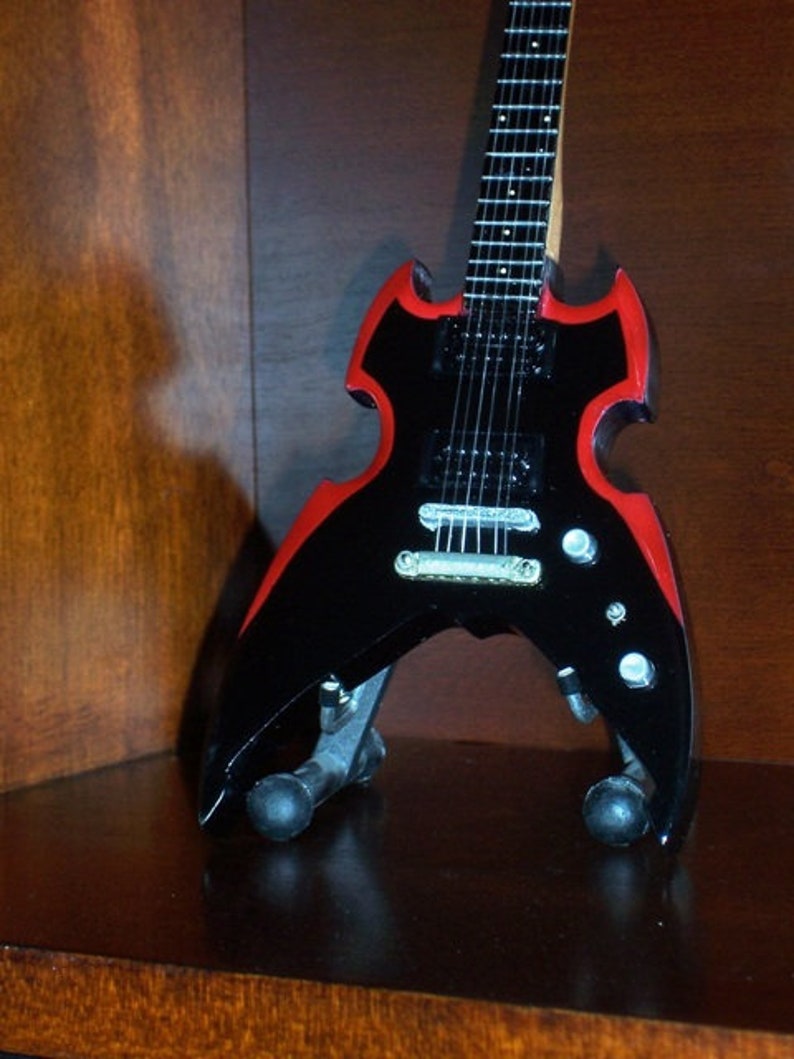 Puede incluir: Una guitarra el&eacute;ctrica negra con detalles rojos. La guitarra tiene una forma &uacute;nica con una pala puntiaguda y un cuerpo que se asemeja a las alas de un murci&eacute;lago. La guitarra tiene una sola pastilla y un puente con una barra de tr&eacute;molo.