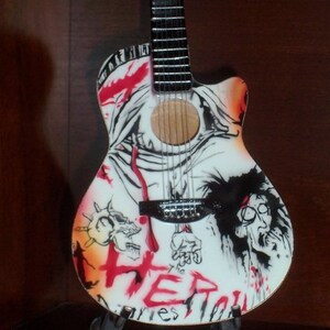 Puede incluir: Una guitarra ac&uacute;stica blanca con un dise&ntilde;o gr&aacute;fico negro y rojo. El dise&ntilde;o presenta una calavera, una figura con una corona de espinas y la palabra "The" seguida de "HER" en rojo. La guitarra est&aacute; en un soporte.