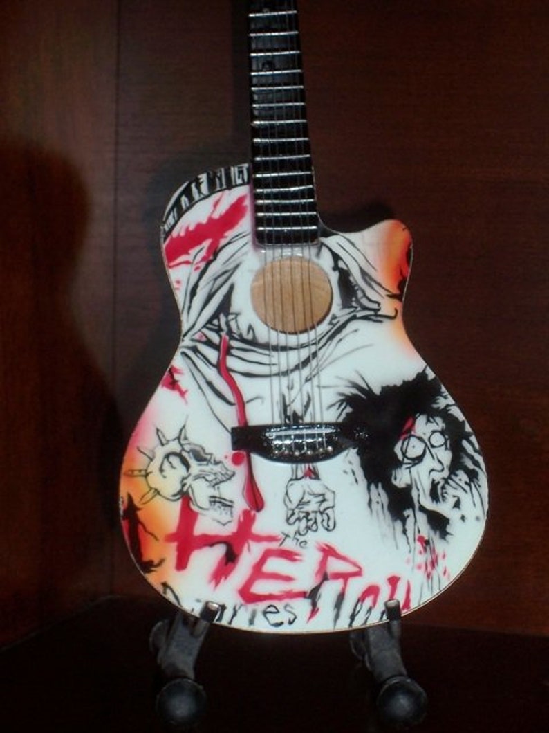 Puede incluir: Una guitarra ac&uacute;stica blanca con un dise&ntilde;o gr&aacute;fico negro y rojo. El dise&ntilde;o presenta una calavera, una figura con una corona de espinas y la palabra "The" seguida de "HER" en rojo. La guitarra est&aacute; en un soporte.