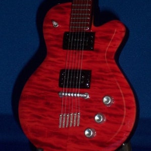 Puede incluir: Una guitarra el&eacute;ctrica roja con una golpeador negro y herrajes cromados. La guitarra tiene una tapa de arce flameado y un cuerpo de caoba.