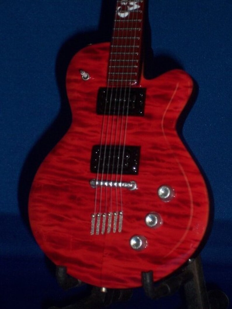Puede incluir: Una guitarra el&eacute;ctrica roja con una golpeador negro y herrajes cromados. La guitarra tiene una tapa de arce flameado y un cuerpo de caoba.