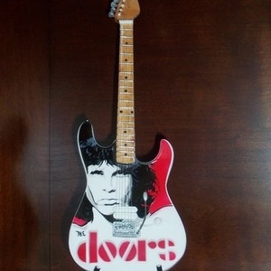 May include: A white electric guitar with a black and red design. The guitar has a portrait of Jim Morrison, the lead singer of The Doors, on the body. The guitar also has the band's name, "The Doors", written in red letters on the body.