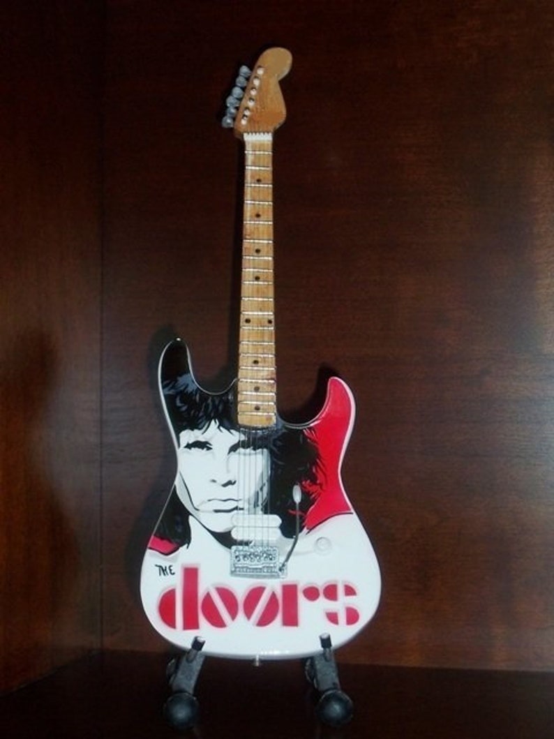 May include: A white electric guitar with a black and red design. The guitar has a portrait of Jim Morrison, the lead singer of The Doors, on the body. The guitar also has the band's name, "The Doors", written in red letters on the body.